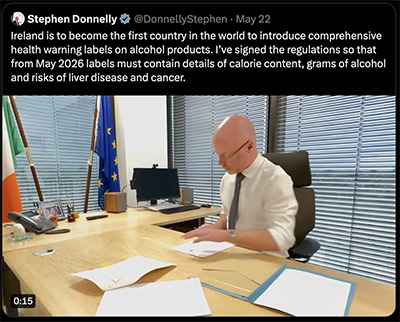 a tweet describing the signing of the first alcohol labeling requirement in Ireland above a screen capture of a bald white man in galsses, white collared shirt, and black tie, sitting at a wood desk, signing papers